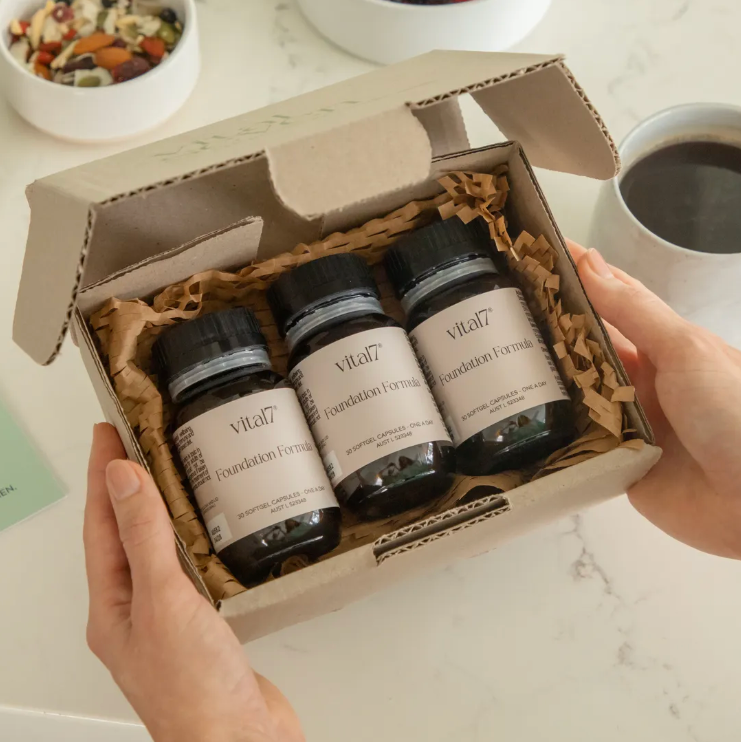 Person holding a box of 'vital seven' products on a kitchen counter with a cup of coffee and snacks.